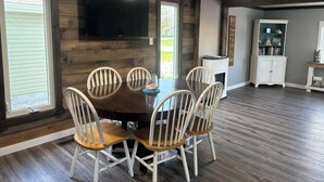 A dining area with a dark wooden table and white chairs, a flat-screen TV mounted on the wall, and a white cabinet with decorative items.