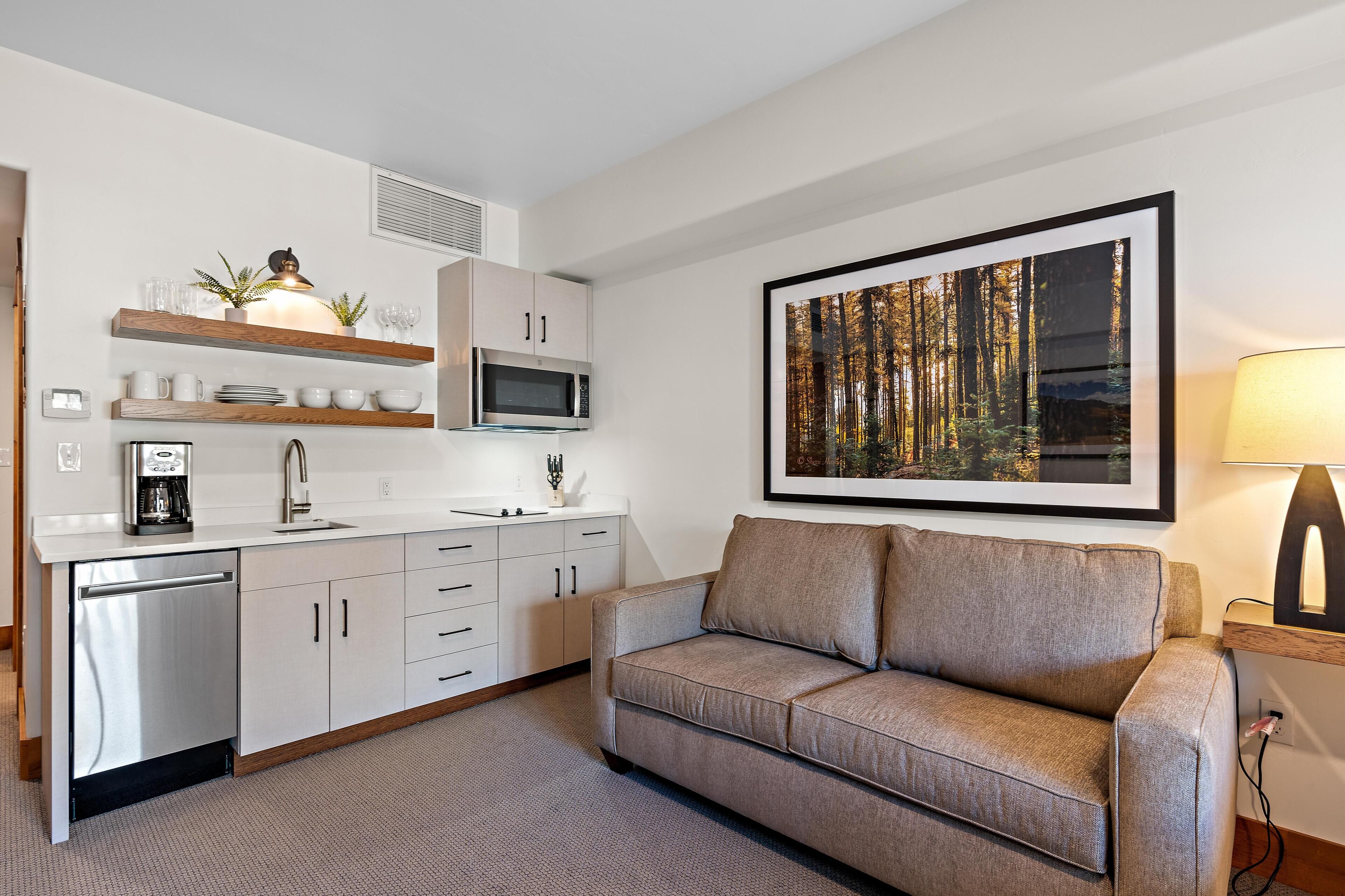 A modern kitchen with white cabinets, a microwave, and a coffee machine. There is a sofa and a framed picture on the wall.