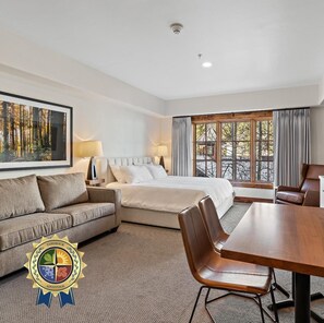 A hotel room with a bed, a sofa, a dining table, and a large window with curtains.