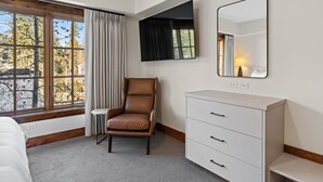 Une chambre d’hôtel avec une grande fenêtre, un fauteuil en cuir marron, une commode blanche et un miroir au mur.