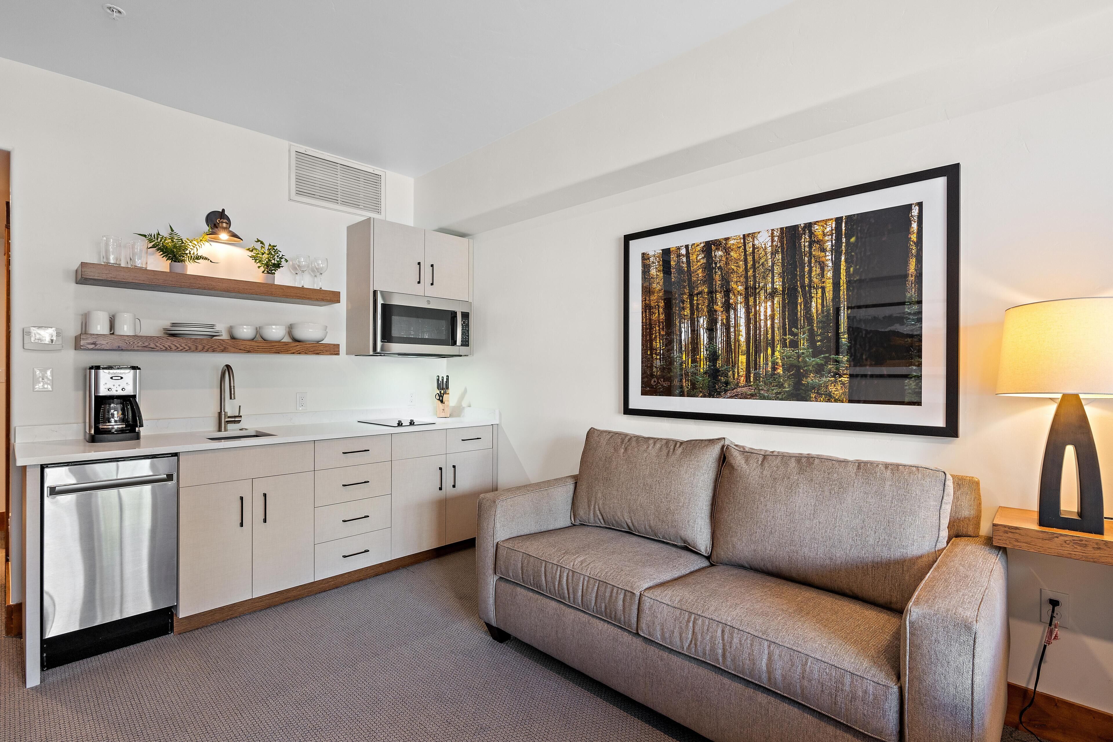 A modern kitchen with a built-in microwave, a coffee machine, and a sink. There is a sofa and a framed picture of a forest on the wall.