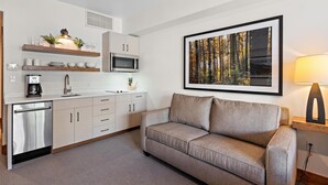 Une cuisine moderne équipée d’un four à micro-ondes encastré, d’une machine à café et d’un évier. On y trouve également un canapé et, au mur, un tableau représentant une forêt.