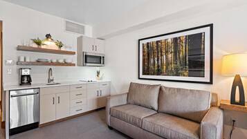 A modern kitchen with a built-in microwave, a coffee machine, and a sink. There is a sofa and a framed picture of a forest on the wall.