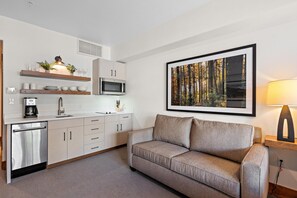 A modern kitchen with a built-in microwave, a coffee machine, and a sink. There is a sofa and a framed picture of a forest on the wall.