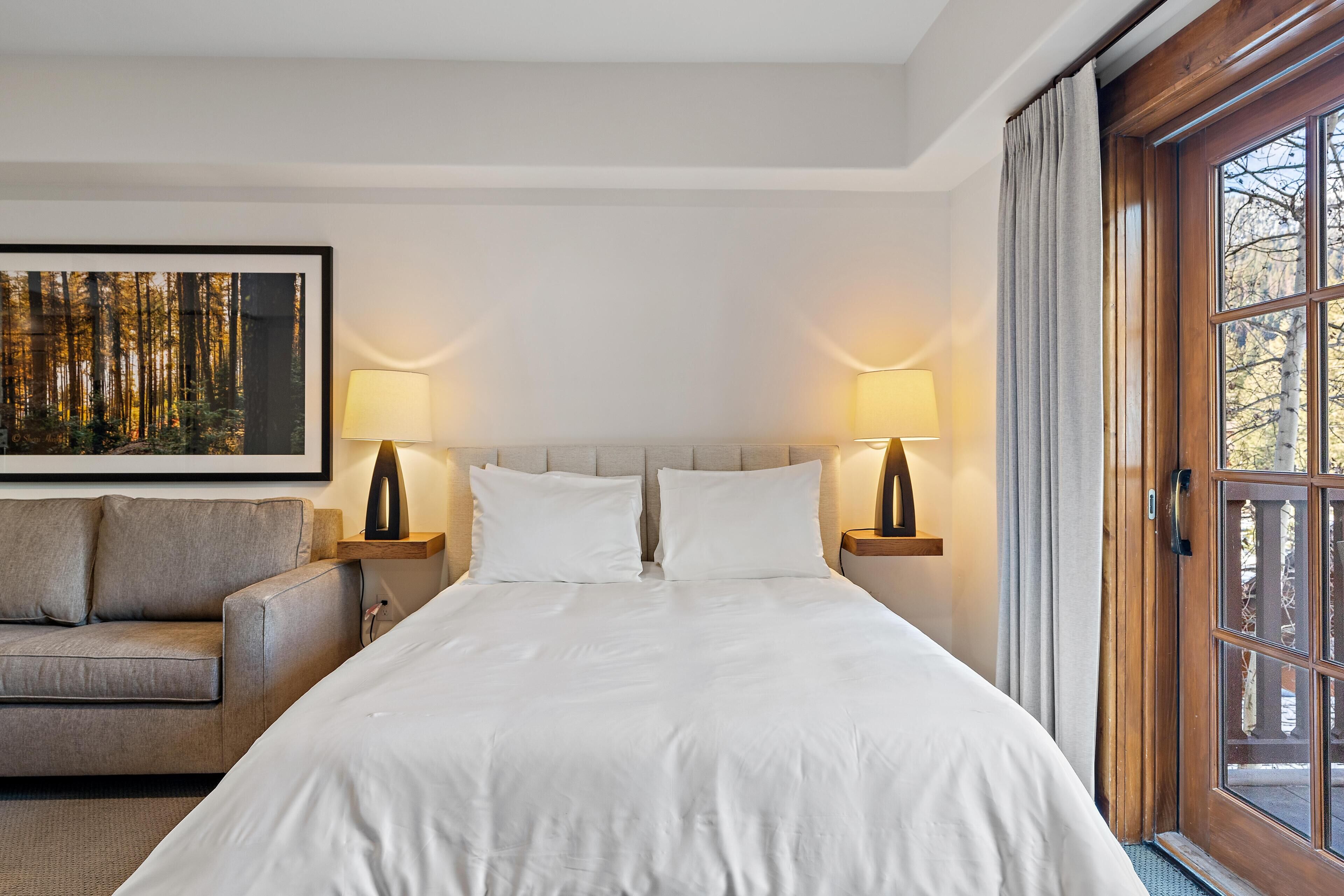 A hotel room with a bed, two bedside lamps, a framed picture on the wall, a sofa, and a wooden door with a view of trees.