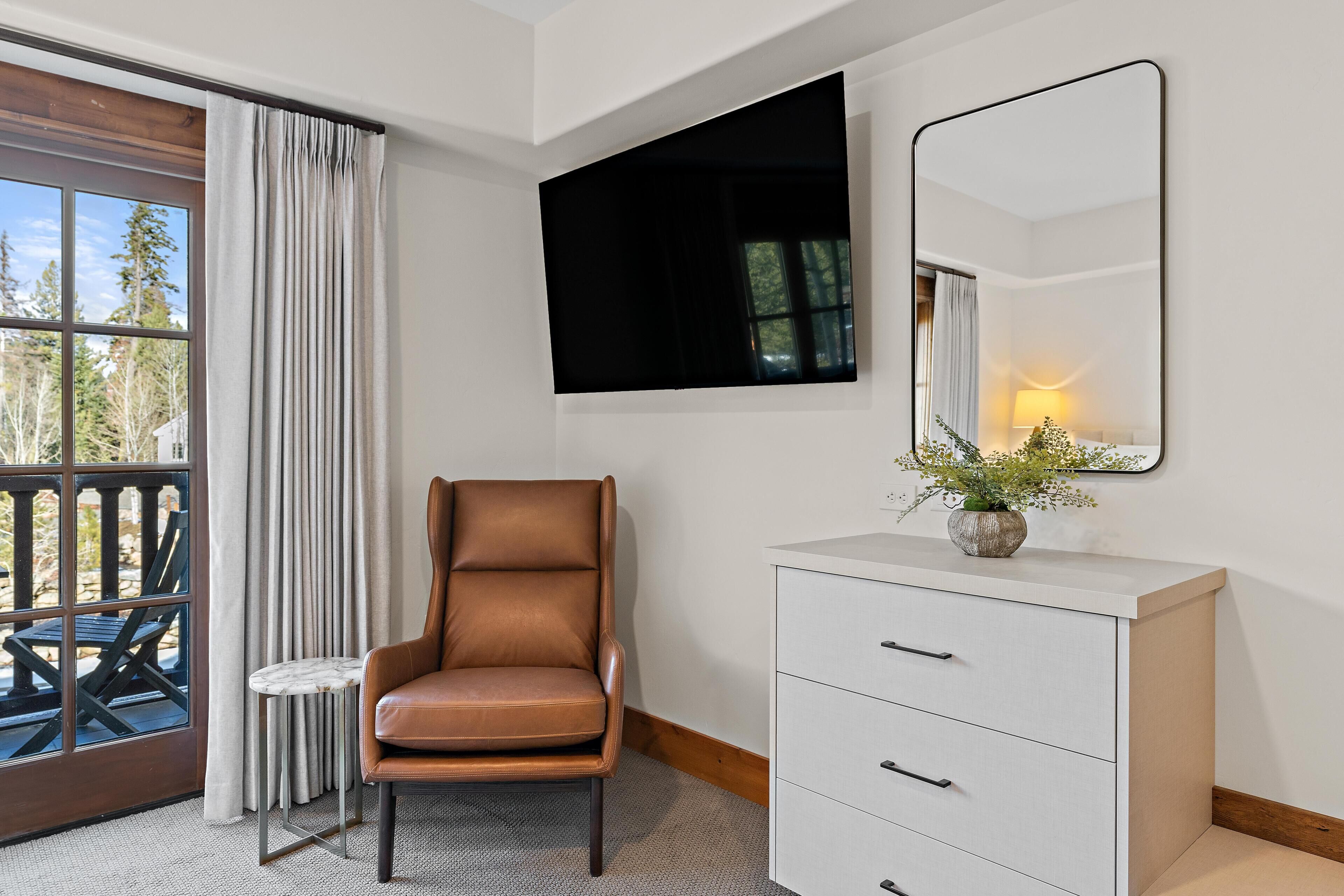 A hotel room with a brown leather chair, a white dresser, a mirror, and a window with curtains.