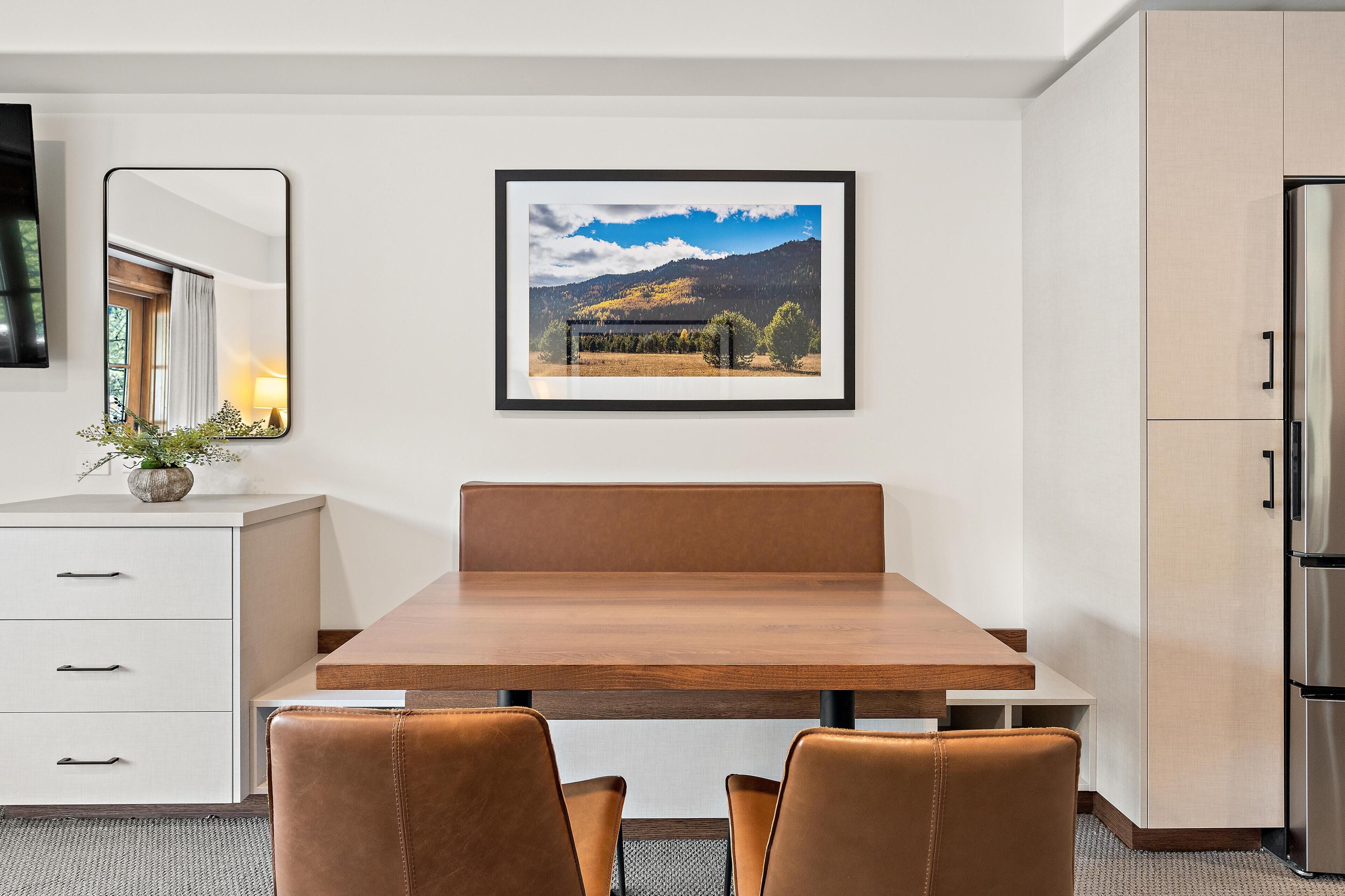 A modern dining area with a wooden table, two leather chairs, a white cabinet with drawers, a framed landscape picture on the wall, and a refrigerator.