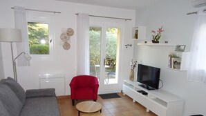 A living room with a grey sofa, a red armchair, two small round tables, a TV on a white stand, and a view through a glass door to the outside.