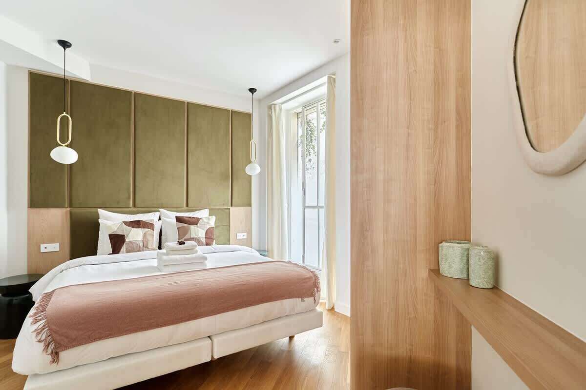Une chambre moderne avec un grand lit, une tête de lit en bois, des luminaires suspendus et une vue sur l’extérieur.