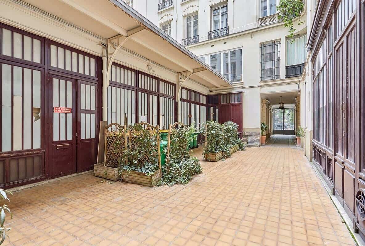 Ein Innenhof mit Fliesenböden, Holz-Pflanzgefässen mit Grünpflanzen und einer Gebäudefassade mit großen Fenstern und einem Schild.