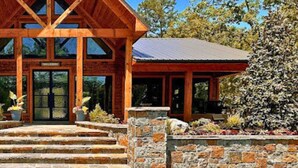 A wooden lodge with a steeply pitched roof, stone steps leading to a glass door, and a stone wall with a flower bed in front. - Game-Filled Retreat for Families and Friends (Broken Bow)