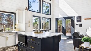 A modern kitchen with a large island, white cabinetry, and black appliances. There is a dining area with a black leather couch and a view of the outdoors. - Game-Filled Retreat for Families and Friends (Broken Bow)
