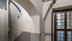 Um corredor com piso de mármore, uma porta com grade de metal e uma parede branca com cano aparente.