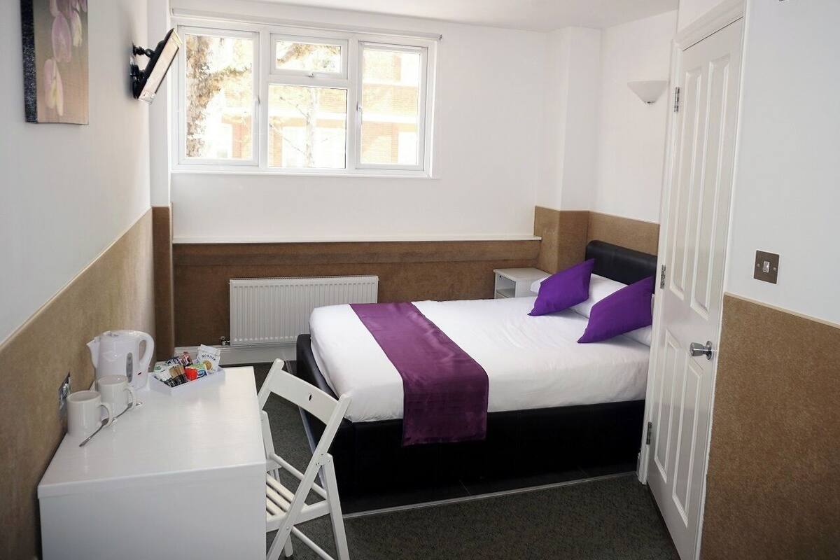 A small, clean room with a bed, a desk, and a chair. There is a window providing natural light, and a radiator under the bed.