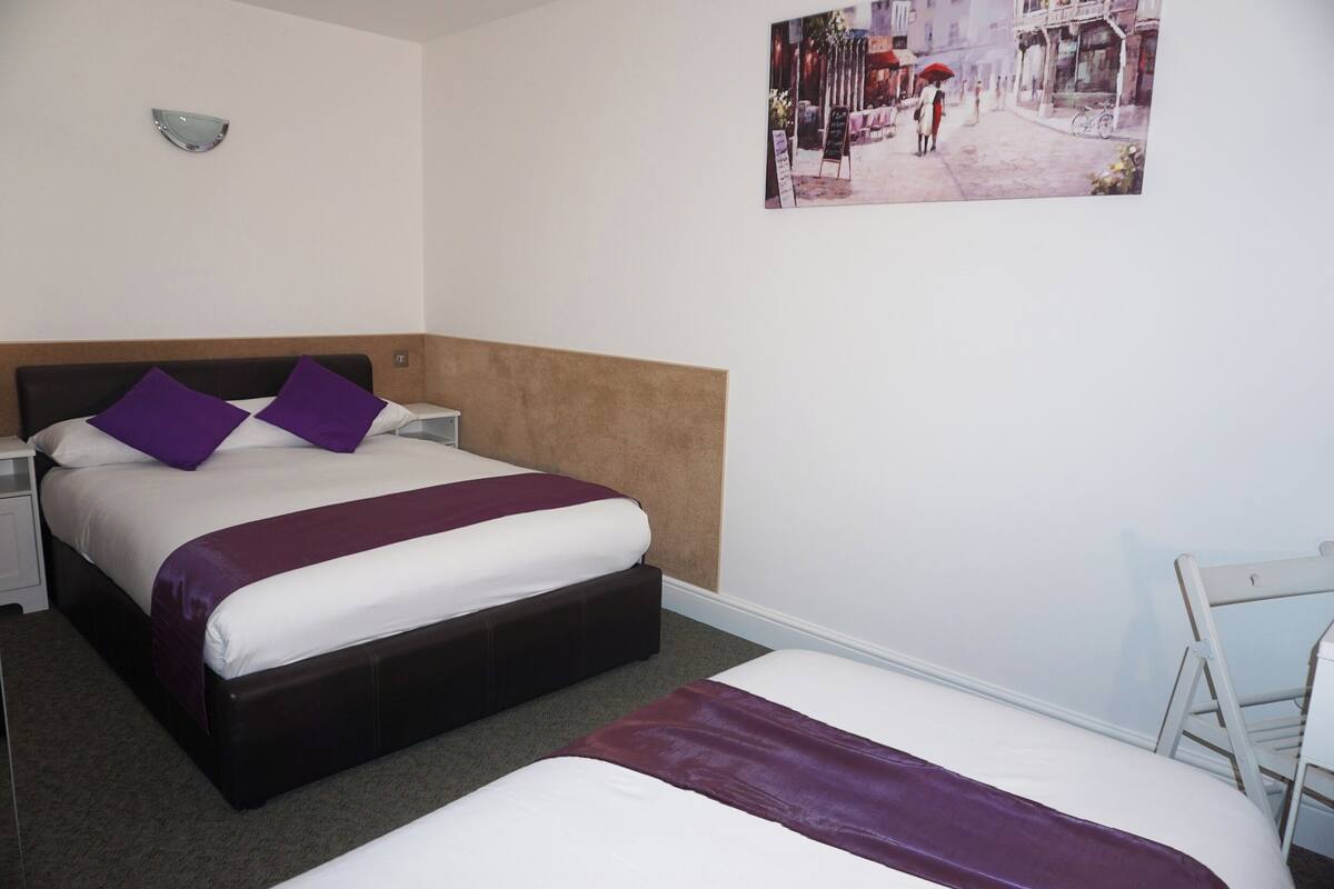 A hotel room with a large bed, a smaller bed, a nightstand, a chair, and a wall-mounted light fixture.