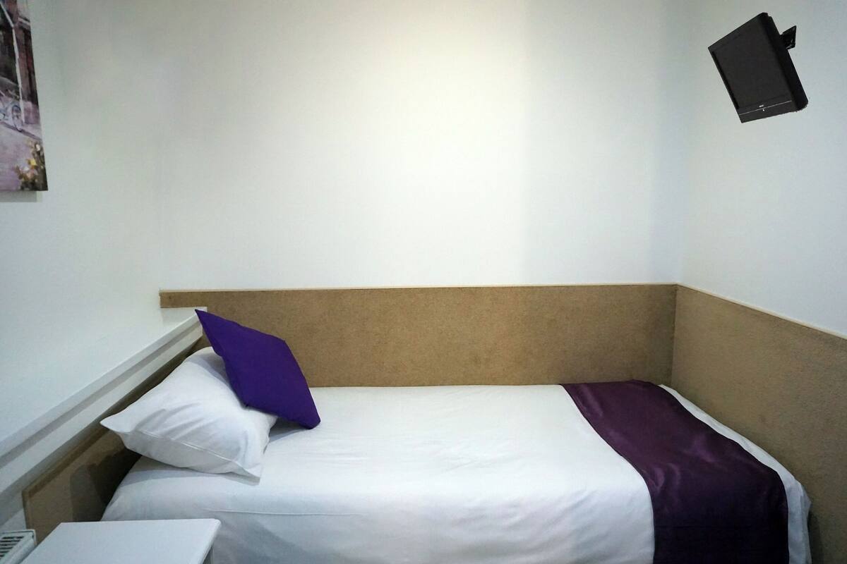 A single bed with white bedding and a purple pillow, a laptop on the left side, and a black object on the wall to the right.