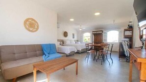A spacious living area with a sofa, a wooden coffee table, and a dining table with chairs. There is a bed with white bedding in the background. - Bella Buzios Apartamentos (Búzios)