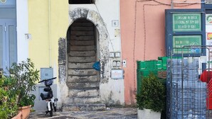 Une cour avec un sol en pierre, des plantes en pot, un scooter et un bâtiment doté d’une porte et d’une enseigne.