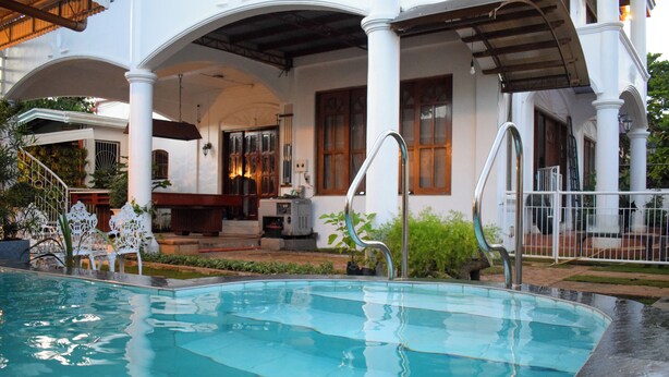 A swimming pool with a covered patio area, a wooden deck, and a view of a building with columns and windows. - Orokasa Guesthouse (Puerto Princesa)