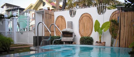 A swimming pool with a stone oven, surrounded by a white wall with wooden doors, and a metal fence.