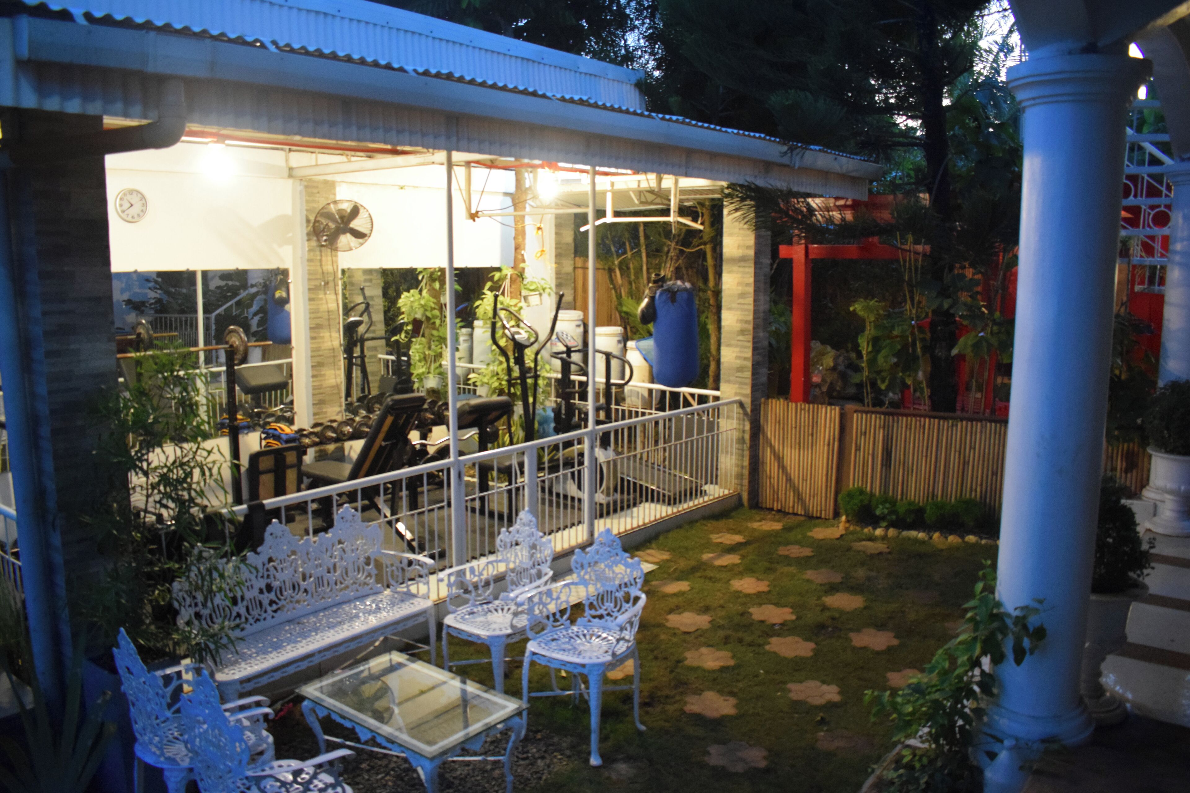 Un patio exterior con mobiliario de metal blanco, una mesa de vidrio y una zona de estar cubierta con un reloj.