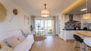 A modern living room with a white sofa, a dining area with a table and chairs, a kitchen with white cabinets and a black countertop, and a balcony with a view of the sea. - Apartamento Lake & SEA (Valencia)