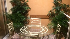 Een tafel met een glazen blad en vier stoelen, elk voorzien van een kleurrijk kussen. Er staan potplanten en een houten bank tegen de muur.