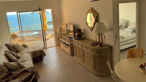 Een ruime woonkamer met een groot raam met uitzicht op de oceaan, een eethoek met een ronde tafel en een gezellige zithoek met een bank.