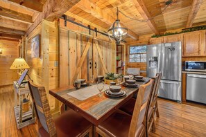 Un interior rústico de madera con una mesa de comedor para cuatro, una araña y un refrigerador moderno de acero inoxidable.