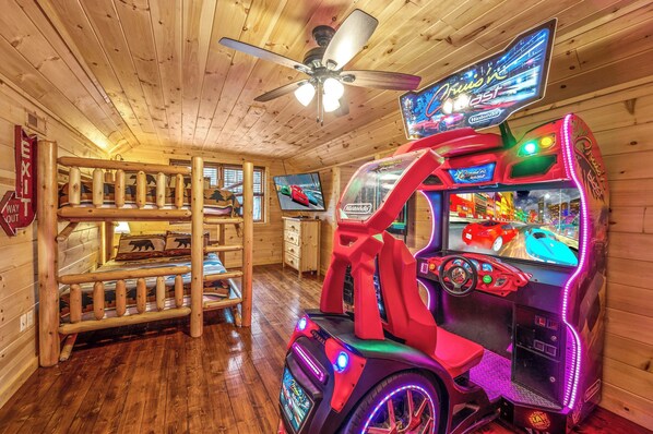 Interior de una cabaña de madera con literas, ventilador de techo y máquina recreativa de carreras.