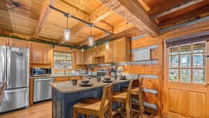 A rustic kitchen with wooden cabinets, a large island with bar stools, stainless steel appliances, and a view through a window. - Secluded Cabin| A+ location <2M DT + Ober| Hot Tub| Firepit| Gameroom (Gatlinburg)