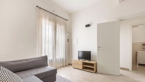 A modern living room with a grey sofa, a flat-screen TV on a wooden stand, a large window with sheer curtains, and a door leading to another room. - 3882 Residenza Virginia by Barbarhouse (Tricase)