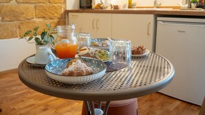 Una mesa de desayuno con diversos alimentos, un vaso de zumo de naranja y una cafetera al fondo.