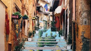 Una calle estrecha con escalones que conducen a un edificio, rodeada de edificios antiguos con balcones y plantas.