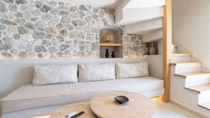 A modern interior with a stone wall, a wooden staircase, a sofa with patterned cushions, and a round table with a remote control. - Alaya Suites (Pyrgos)