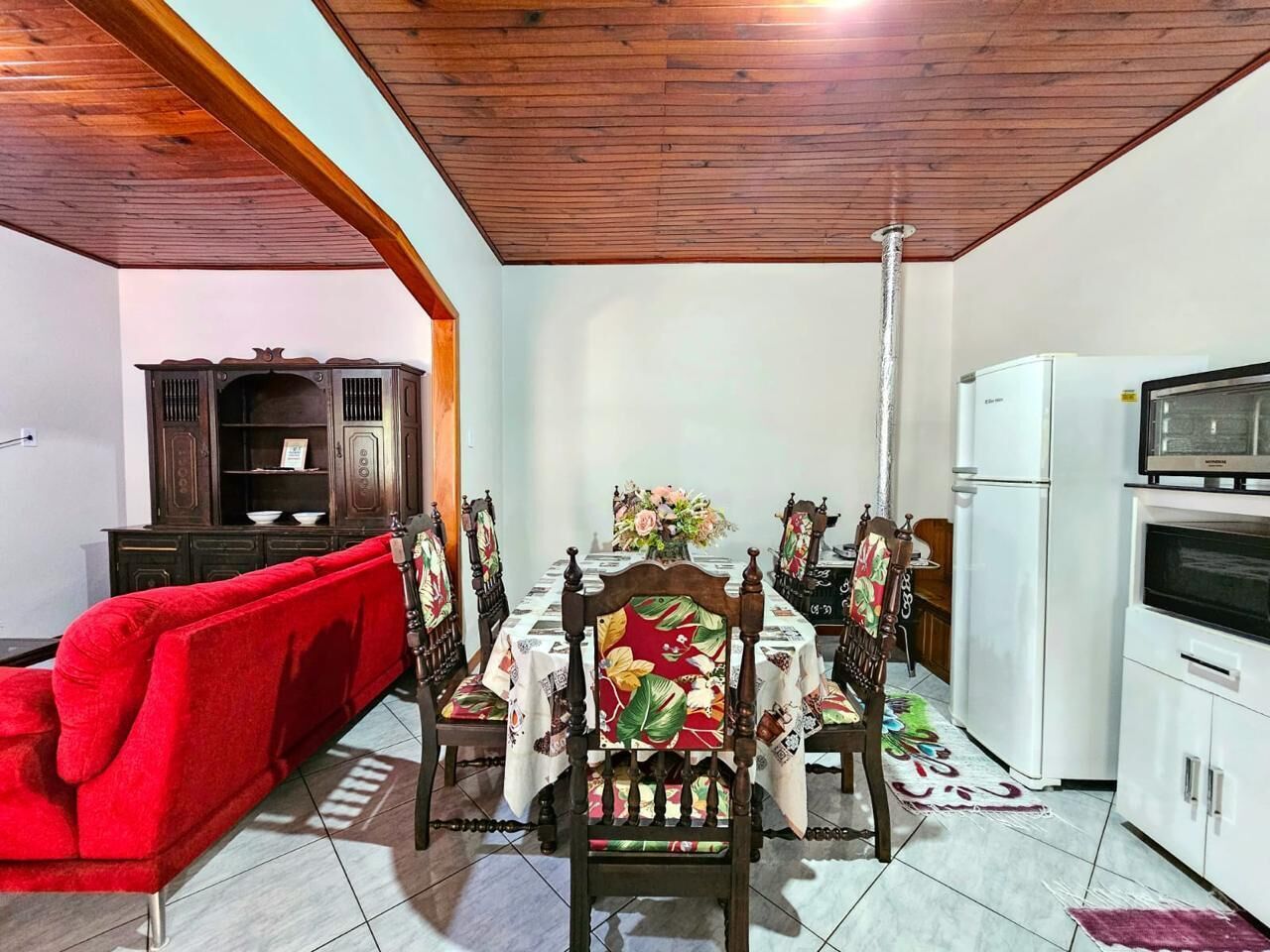 A dining area with a wooden table set for four, surrounded by chairs with floral upholstery. A red sofa is positioned to the left, and a white refrigerator is visible to the right.