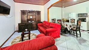 Una sala de estar con techo de madera, un sofá rojo, un mueble de madera, una mesa de comedor con sillas y una zona de cocina con microondas.