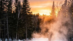 Una piscina rectangular con dos personas sentadas al final, rodeada de nieve y árboles, y una puesta de sol al fondo.