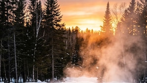 A rectangular pool with two people sitting at the end, surrounded by snow and trees, with a sunset in the background. - Nomi - King Suite (314) (Harcourt)