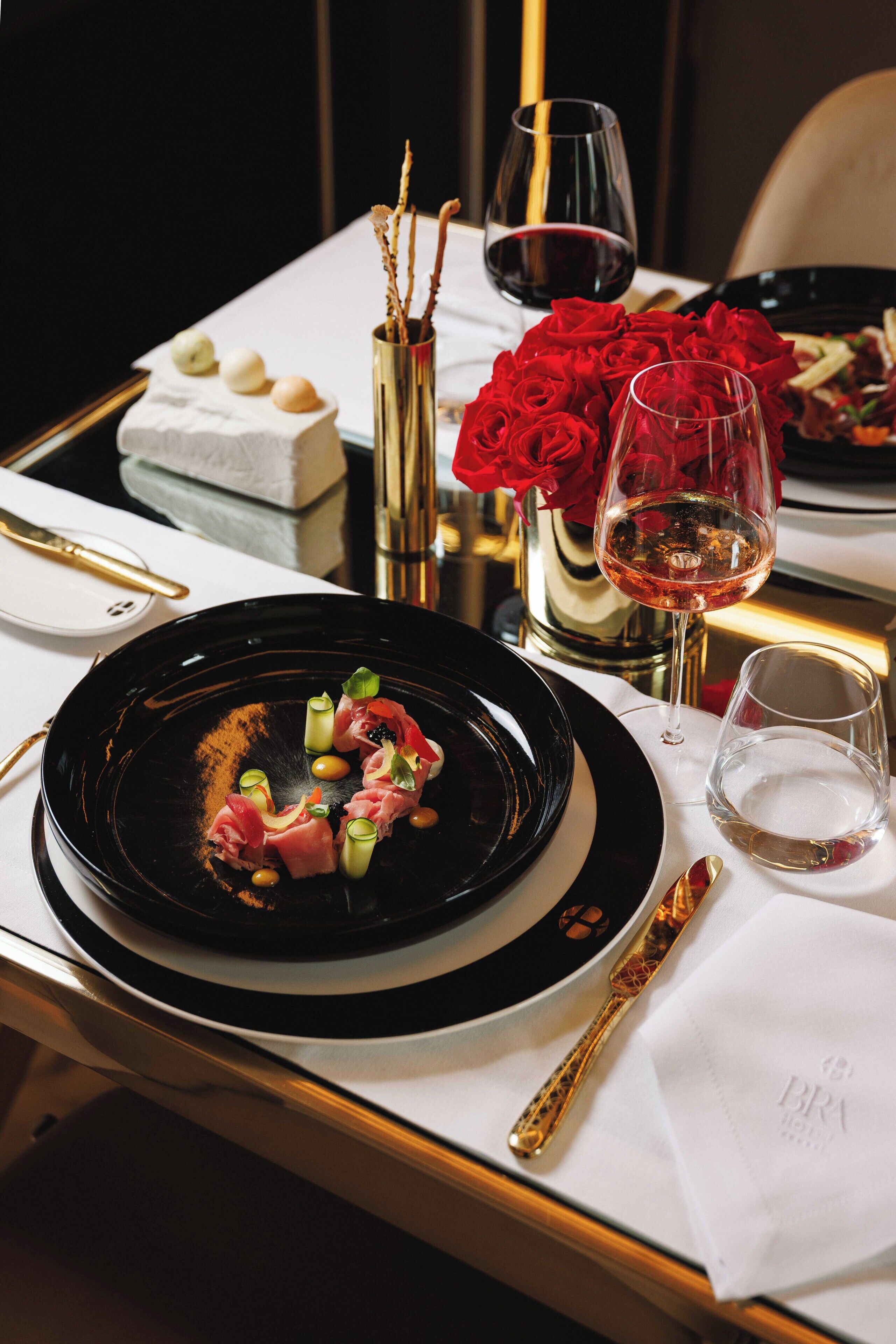 Une table à manger avec une assiette noire, un verre de vin rouge, un vase rempli de roses rouges et un vase de couleur or orné de branches.