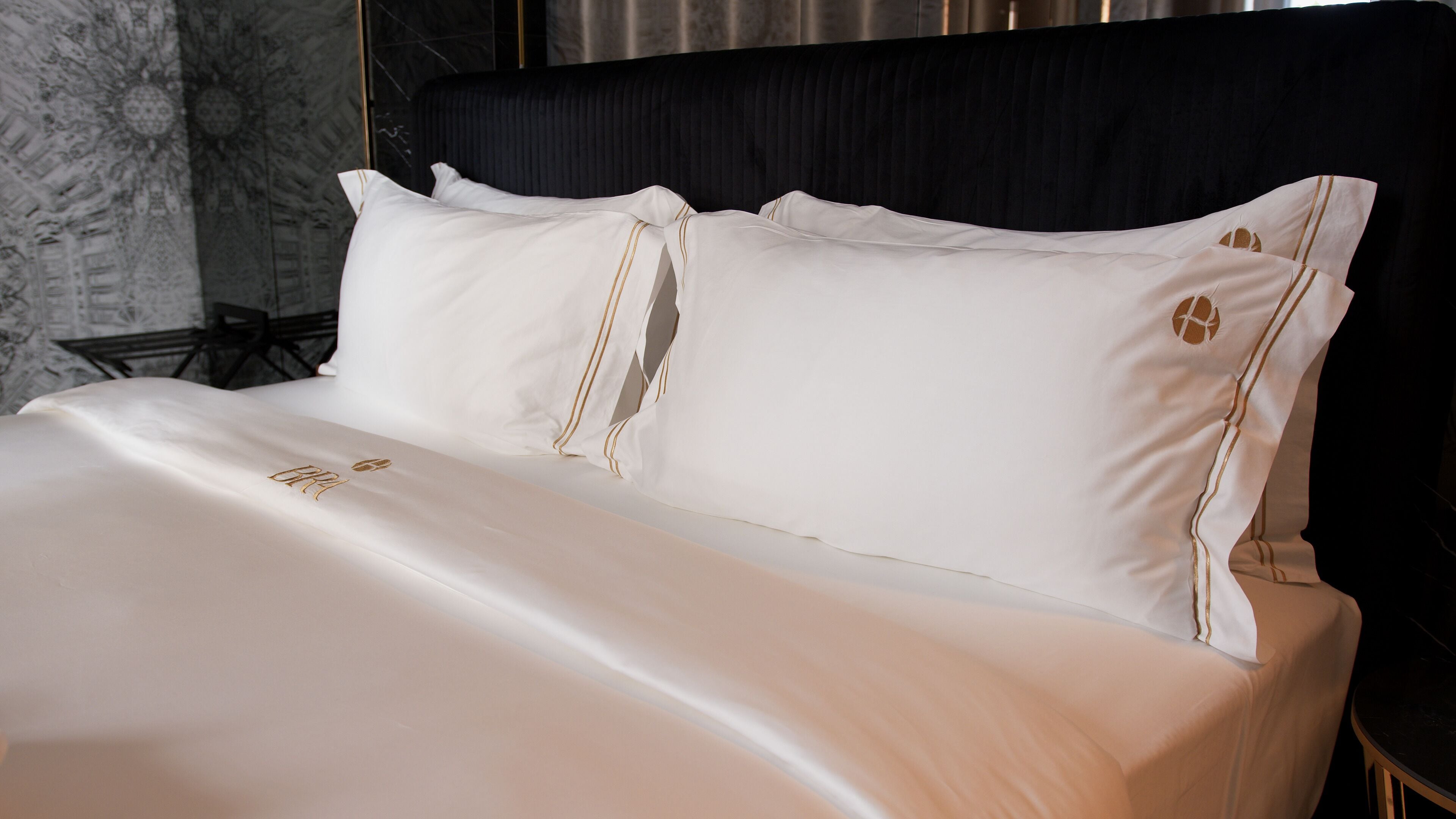 A neatly made bed with white linens and gold accents, a dark headboard, and a marble-patterned wall.