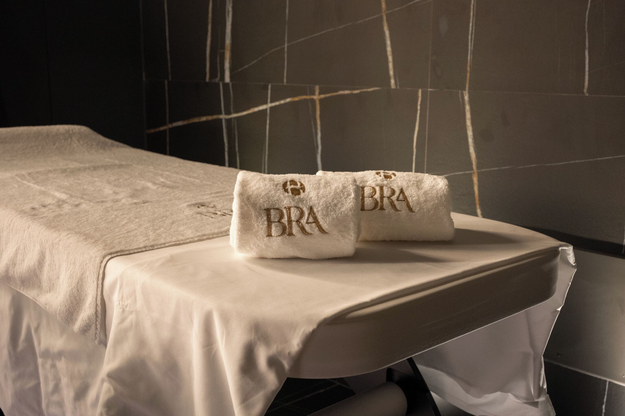 A neatly made bed with a white bedspread and two towels with 'BRA' embroidered on them.