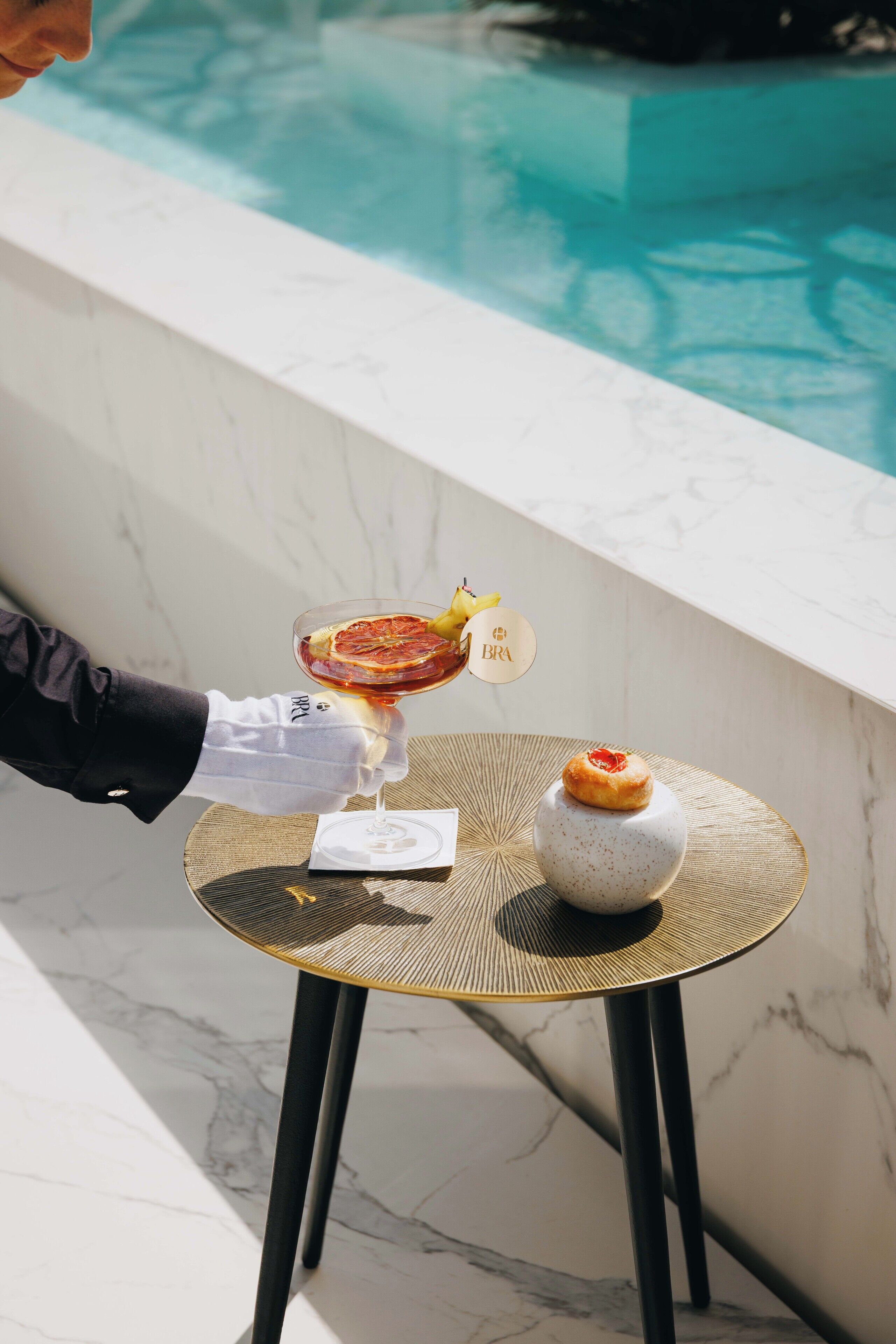 Una persona sosteniendo una copa de cóctel con una guarnición, un postre sobre una mesita y una piscina al fondo.