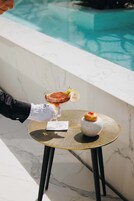 Una persona sosteniendo una copa de cóctel con una guarnición, un postre sobre una mesita y una piscina al fondo.