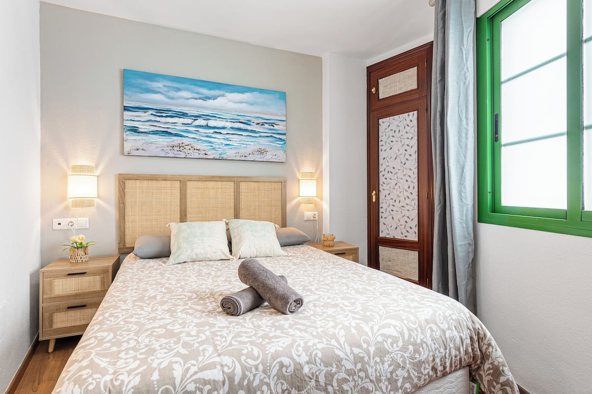 Een keurig opgemaakt bed met een beddengoed met een patroon, twee bedlampjes, een nachtkastje met een plant, en een schilderij van de oceaan aan de muur.