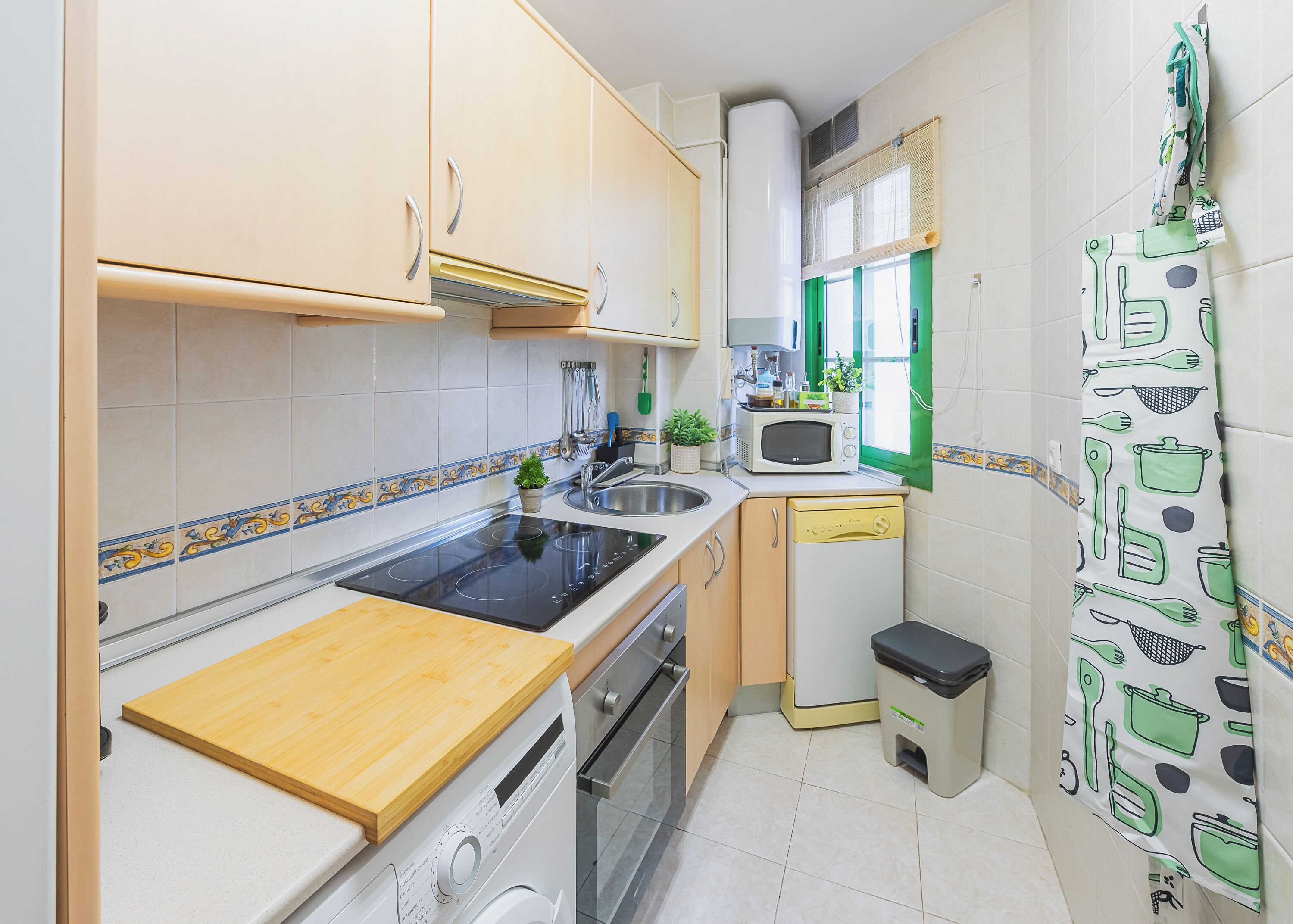 A compact kitchen with a stove, sink, microwave, and a small trash bin.