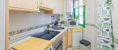 A compact kitchen with a stove, sink, microwave, and a small trash bin.