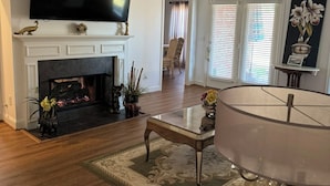 Uma sala de estar ampla, com lareira, TV de tela plana, ventilador de teto e uma grande janela com persianas.