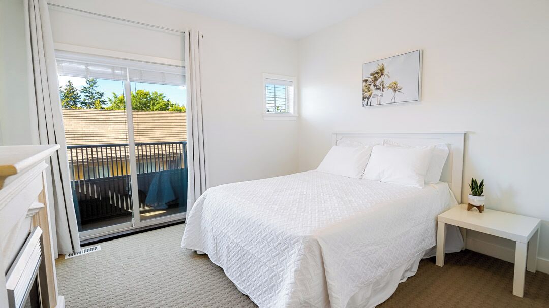 A bedroom with a large bed, a nightstand, a small table, a window, and a balcony with a view of greenery.