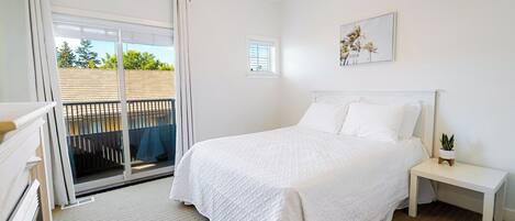 A bedroom with a large bed, a nightstand, a small table, a window, and a balcony with a view of greenery.